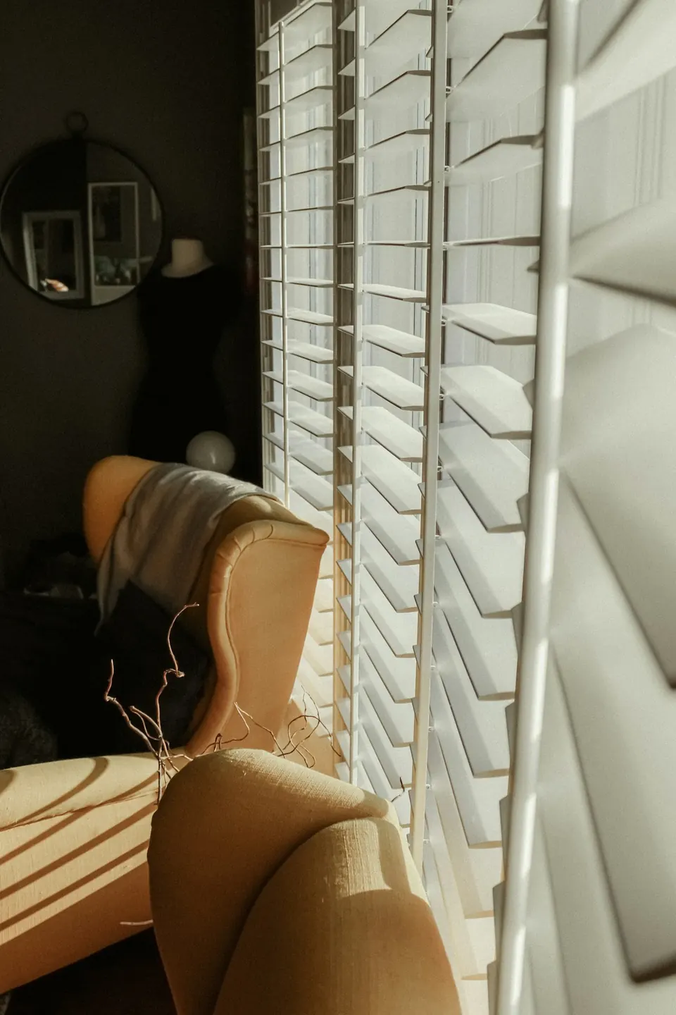 Sunlight through white louvered window shutters beside mustard-yellow armchairs